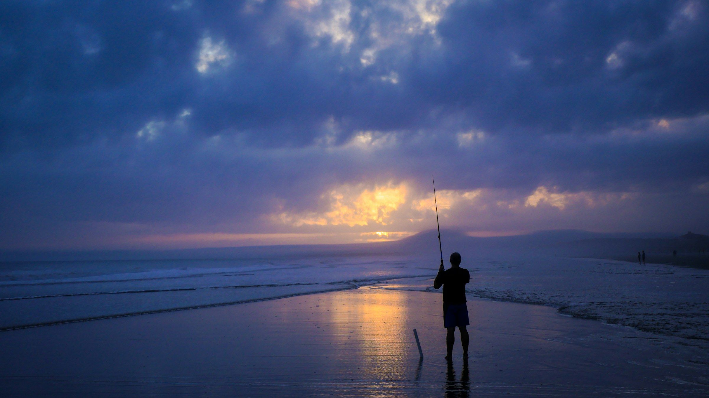 Un pescatore in piedi sulla battigia all’imbrunire, con la canna in mano, sotto un cielo di nuvole viola e un ultimo chiarore sull’acqua.