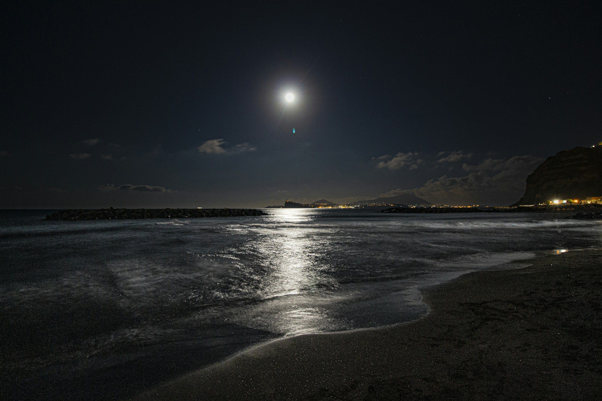 La luna piena sul mare di notte, con il riflesso sulla battigia e la costa illuminata in lontananza.