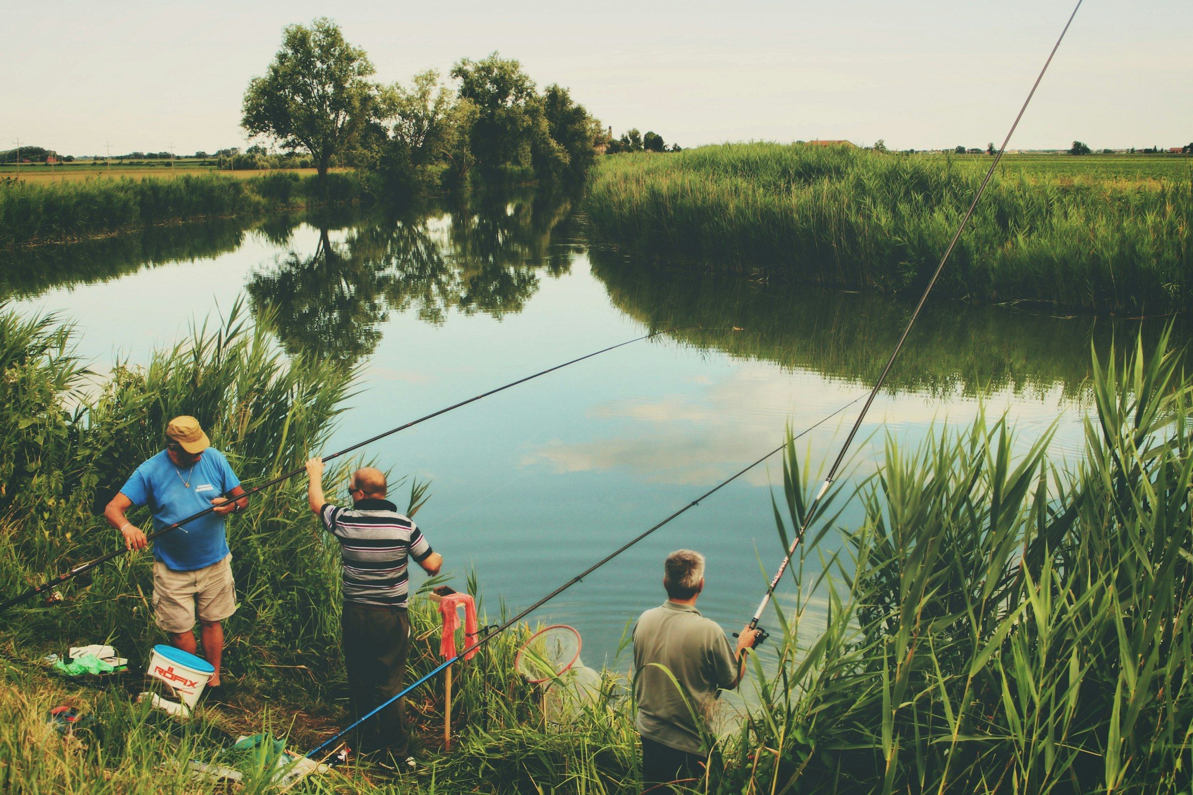 Tre pescatori con le canne lunghe sulla riva di un canale tra i canneti.