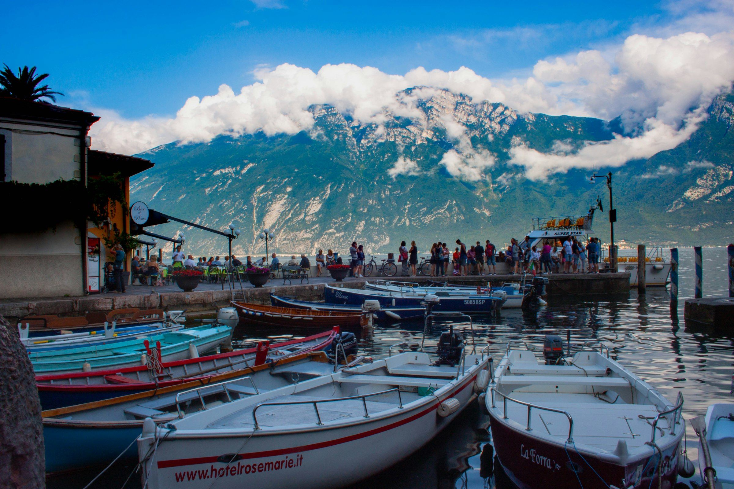 Barche da pesca ormeggiate in un porticciolo del lago di Garda, con le montagne tra le nuvole sullo sfondo.