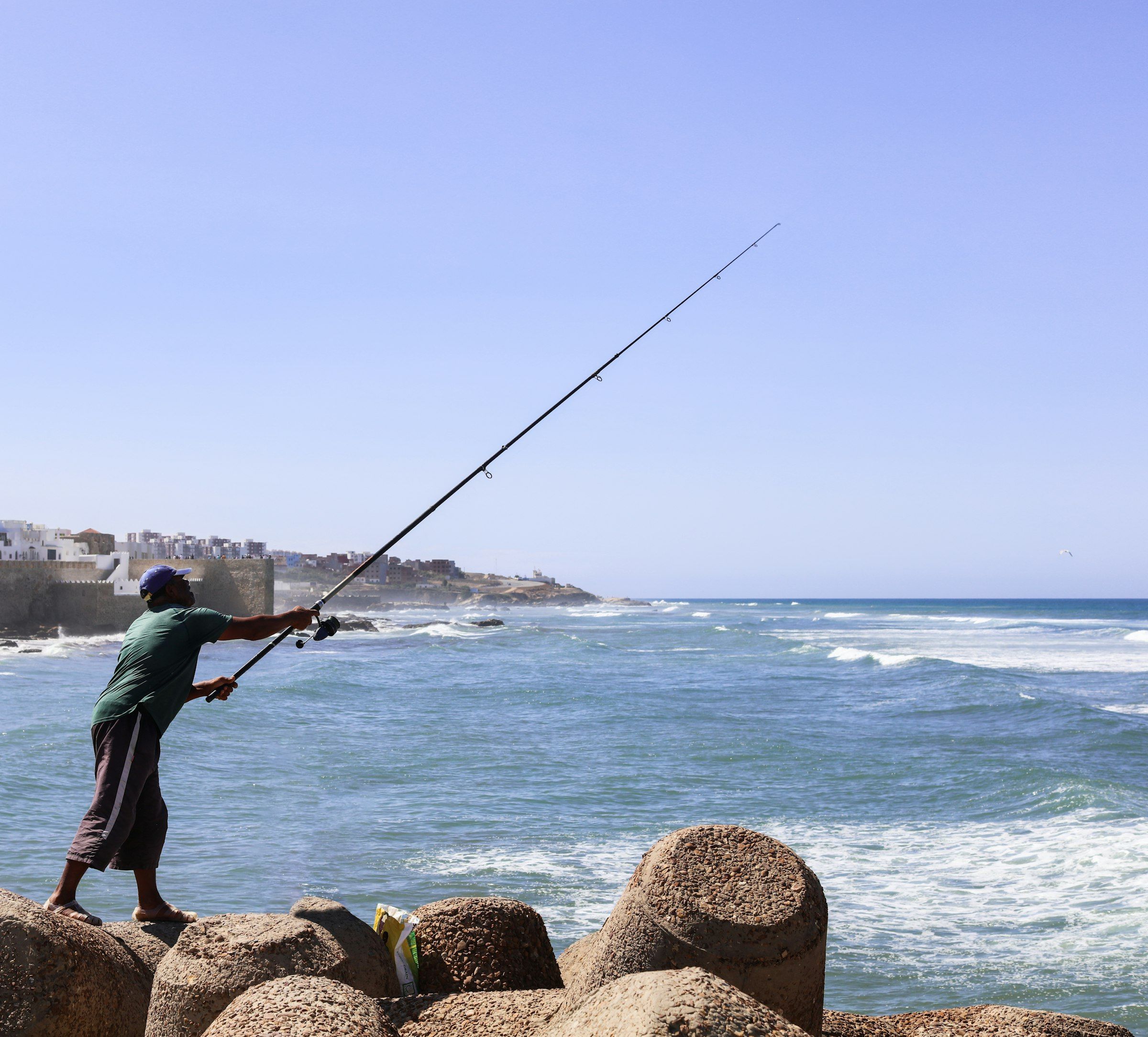 Un pescatore lancia da una scogliera di massi frangiflutti verso il mare aperto, con la canna piegata nel lancio.