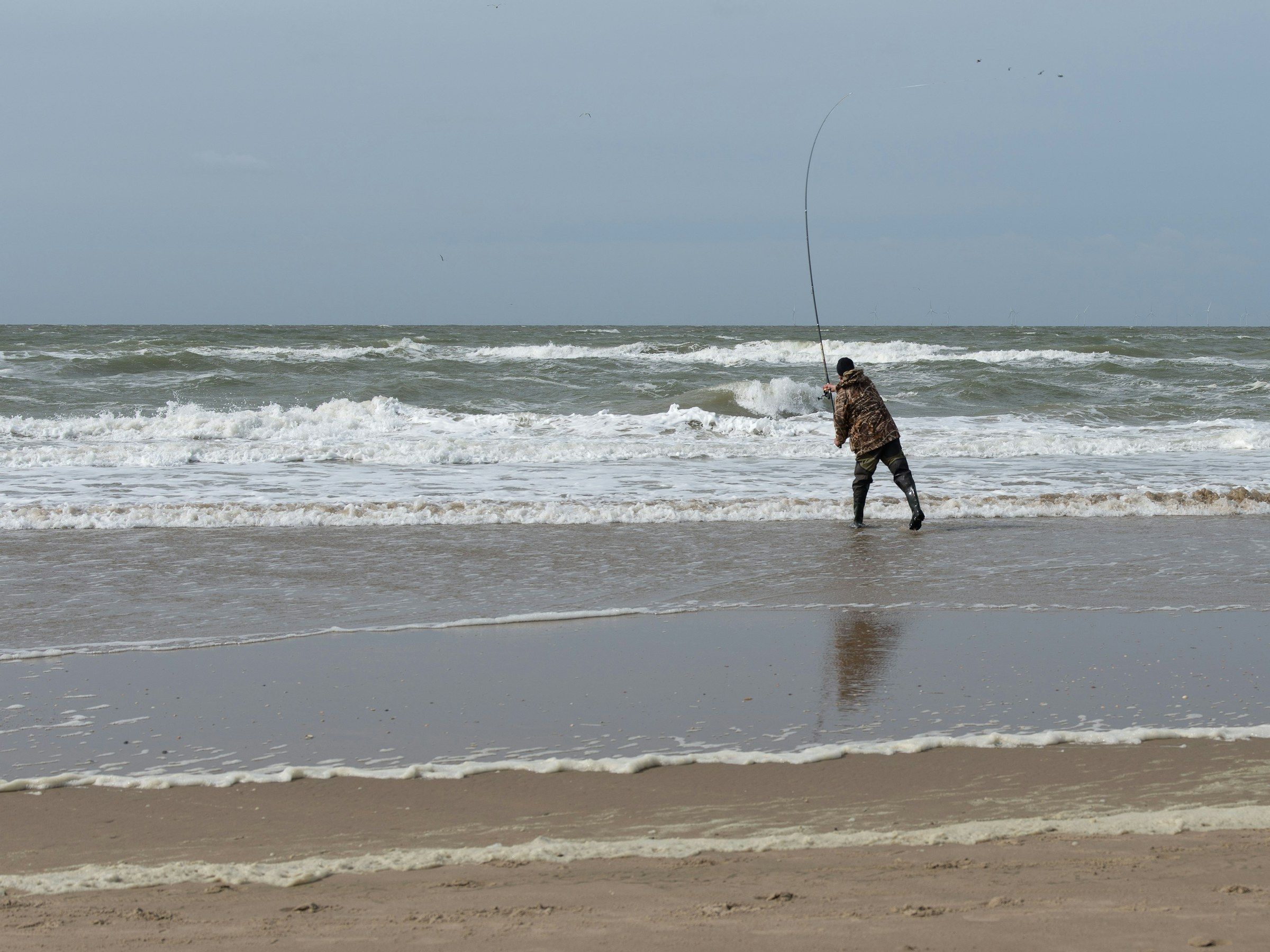 Un pescatore a surfcasting sulla battigia con il mare mosso e le onde che frangono.